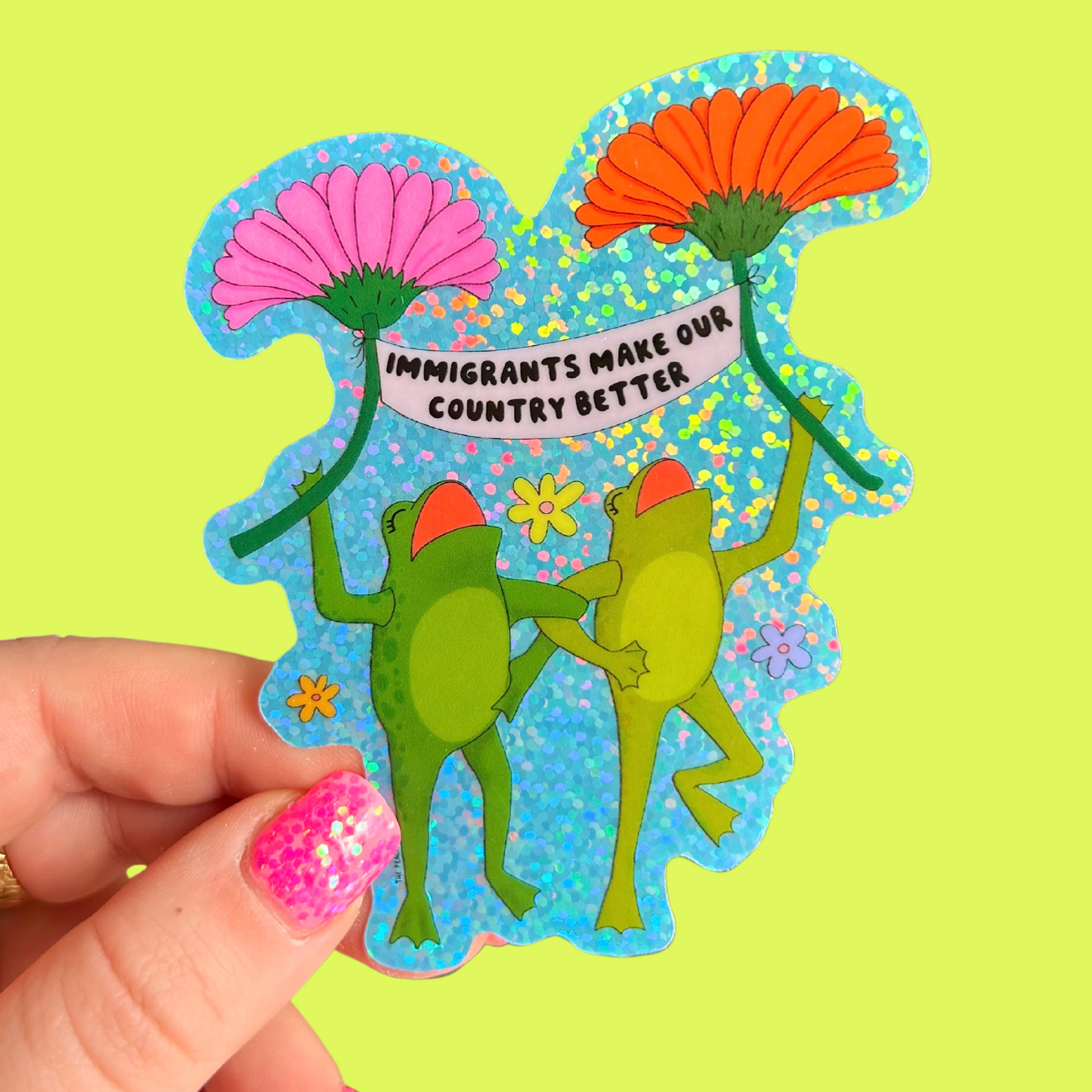 Colorful sticker with frogs and flowers held by a hand on a green background