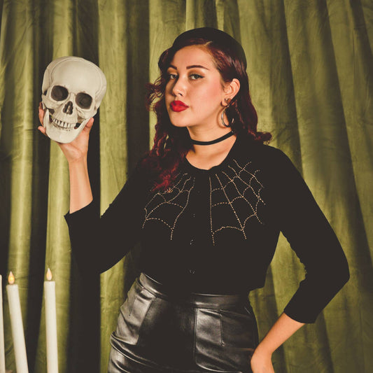 Woman in a black outfit with spider web design holding a skull, standing behind a table with candles and fruit against a green curtain background.
