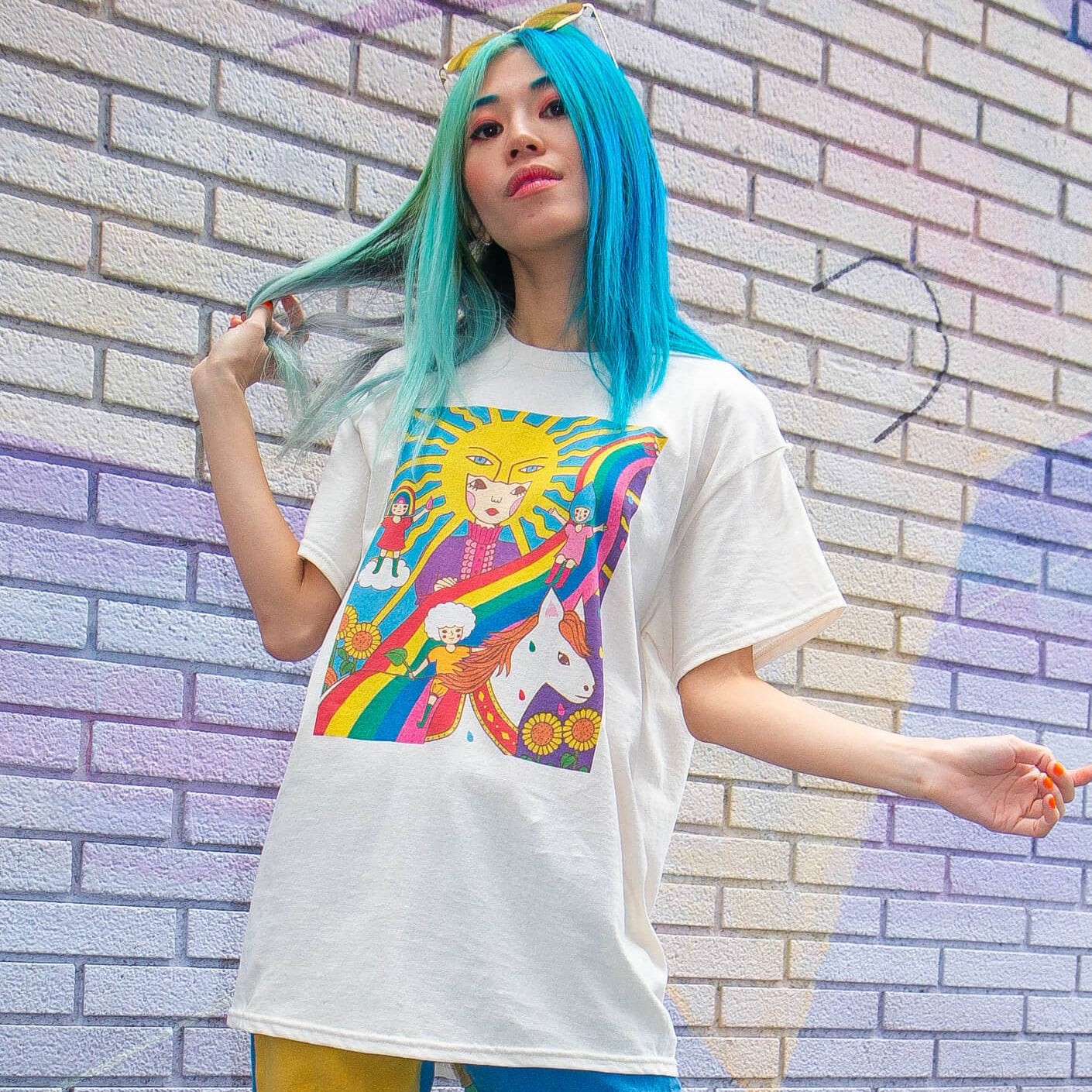 Person with blue hair wearing a white t-shirt with a colorful graphic design against a brick wall.